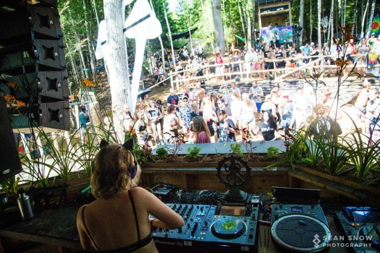 Vue depuis la cabine du DJ sur une foule qui danse en plein jour dans un amphithéâtre forestier bordé de sacs de sable