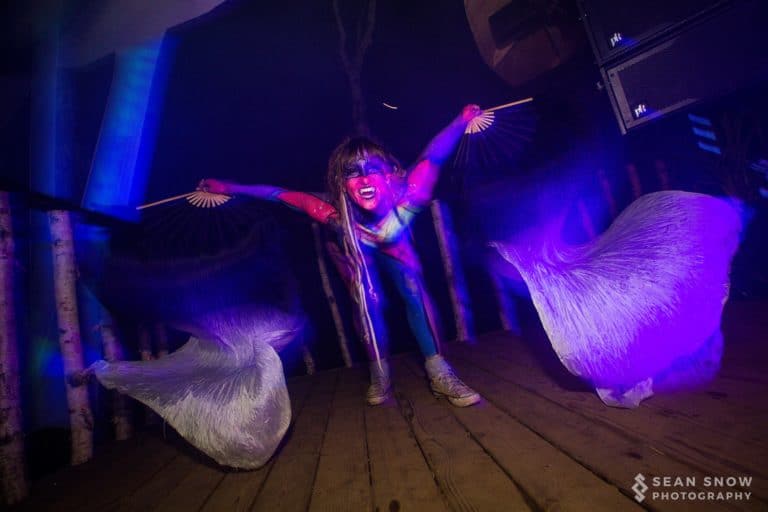 Sous une lumière violette, une personne au maquillage fluo déploie des voiles de soie sur une scène en bois, près de bouleaux