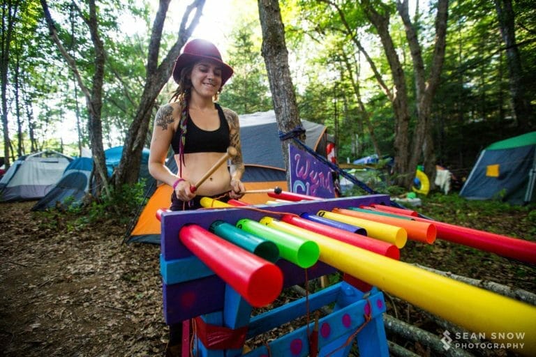 Dans un camping boisé, une personne au chapeau rouge joue d'un xylophone en tubes multicolores avec des mailloches