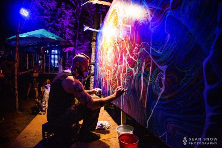 Peinture en direct la nuit, sous lumière noire, avec des lignes blanches tracées sur une toile aux tourbillons arc-en-ciel