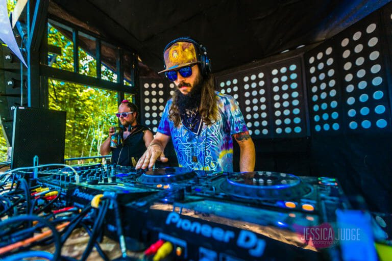 DJ avec casque d'écoute et chemise multicolore aux platines Pioneer, sur une scène de jour en forêt aux panneaux noirs à pois