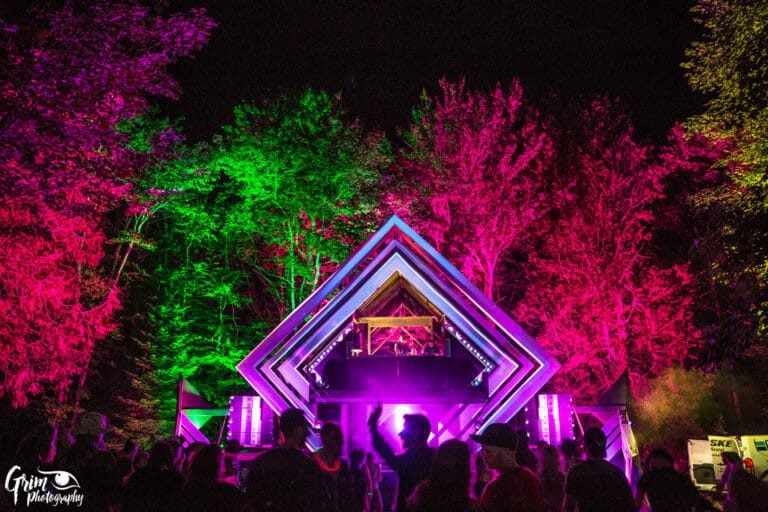 Foule qui danse la nuit devant une scène en losanges emboîtés illuminée de violet, sous des arbres éclairés magenta et vert