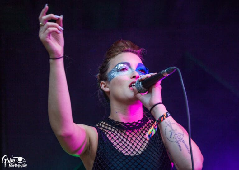 Artiste au maquillage bleu scintillant qui chante au micro sur scène, un bras levé, sous un éclairage rose et violet