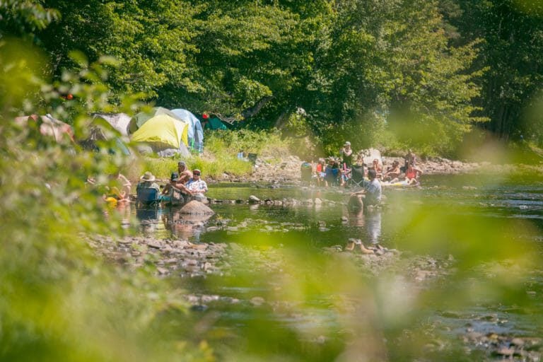Personnes détendues sur des chaises de camping dans une rivière peu profonde et rocailleuse, près des tentes, en plein soleil
