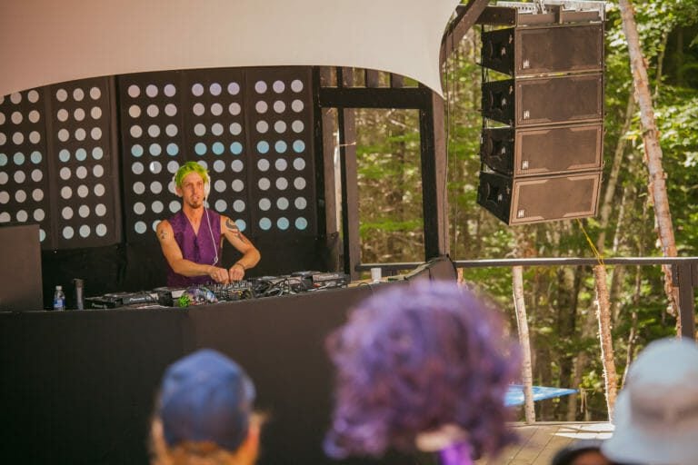 DJ aux cheveux vert vif qui mixe sous l'auvent d'une scène en forêt, de jour, près des haut-parleurs suspendus, public devant