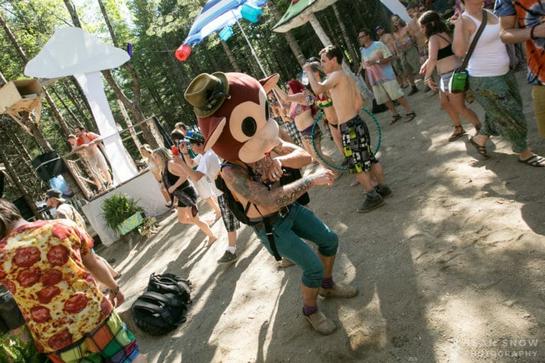 Une personne coiffée d'une grosse tête d'animal de dessin animé danse avec la foule en plein jour, sur la piste en terre