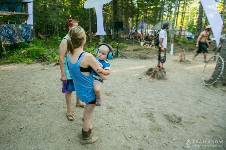 Bébé portant des protecteurs d'oreilles bleus dans les bras d'un adulte, sur une piste de danse ensoleillée en forêt