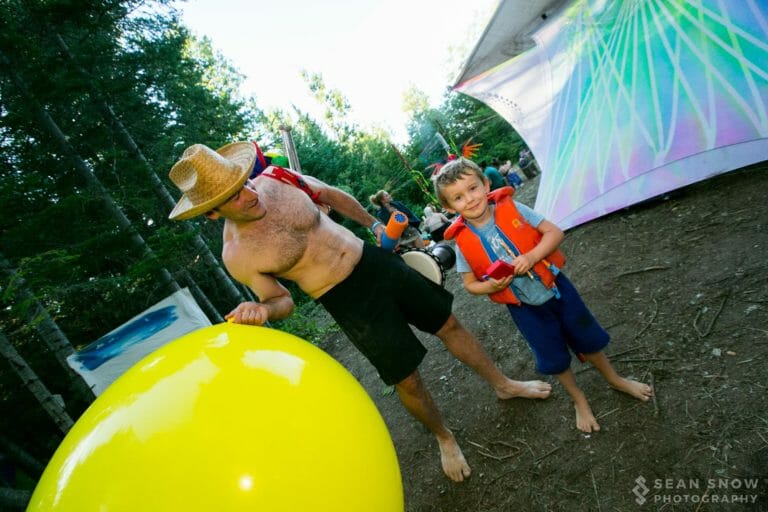 Enfant souriant en gilet de sauvetage près d'un percussionniste au chapeau de paille et d'un gros ballon jaune