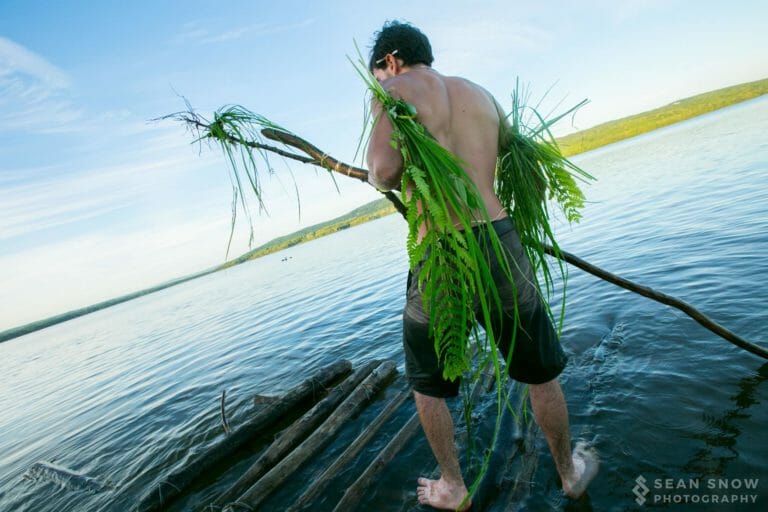 Une personne torse nu couverte de fougères et de joncs monte sur un radeau de rondins du lac, une branche à la main