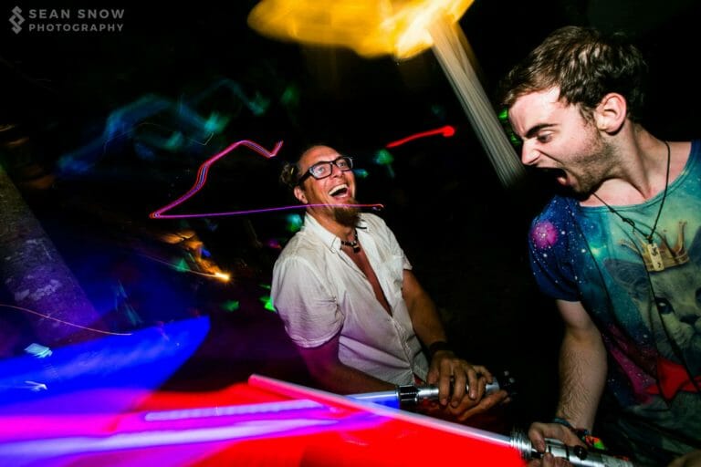 Deux festivaliers rient et crient en croisant des sabres laser jouets, entourés de traînées de lumière colorées la nuit