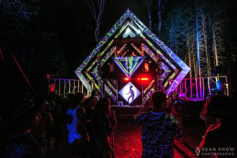 Foule devant une scène en losange dans la forêt, la nuit, sous des projections vertes et violettes et une silhouette dansante