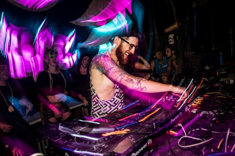 DJ aux bras tatoués, sourire aux lèvres, aux platines parmi des traînées de lumière rose et bleue, public juste derrière