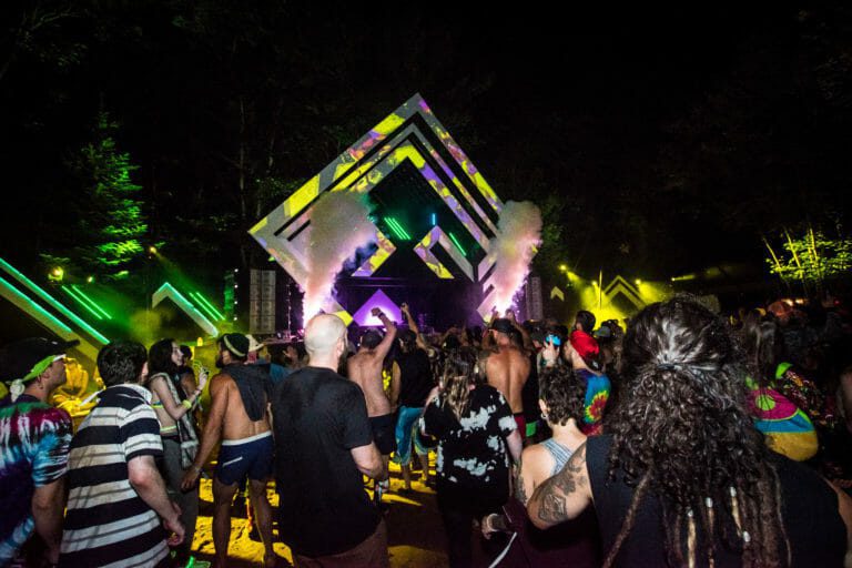 Foule devant une scène lumineuse en losanges superposés, jets de CO2 blancs et lasers verts dans la forêt, la nuit