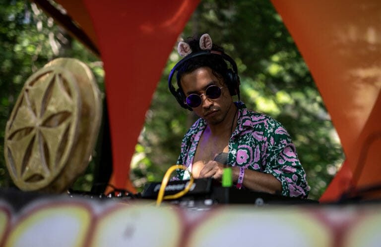 DJ en lunettes rondes, casque et oreilles en peluche, jouant un set de jour sous des voiles d'ombrage orange