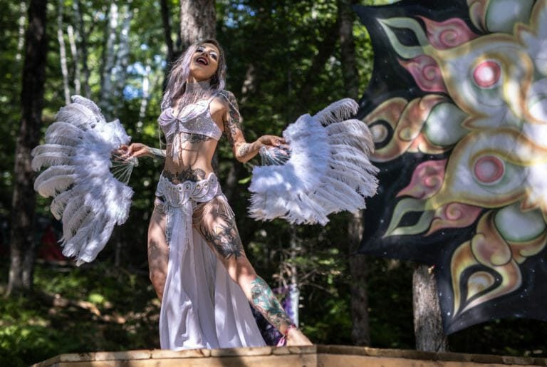 Artiste en blanc, tatouages visibles, dansant avec des éventails de plumes sur une scène de bois en forêt ensoleillée
