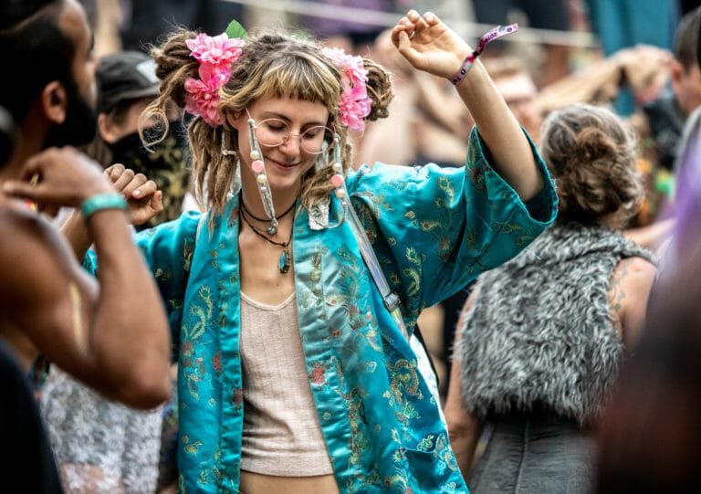 Personne souriante qui danse en veste kimono turquoise à dragons, fleurs roses aux cheveux, bras levé dans la foule le jour