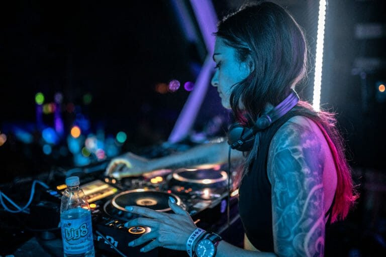DJ au bras tatoué, casque au cou, aux platines sous une lumière bleue et rose, lumières du site floues à l'arrière-plan