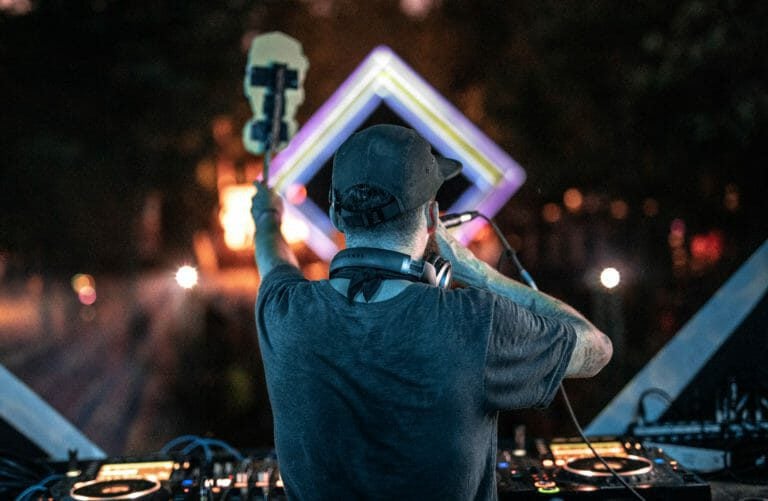 DJ de dos aux platines, brandissant un totem et un micro vers un cadre lumineux en losange, la nuit