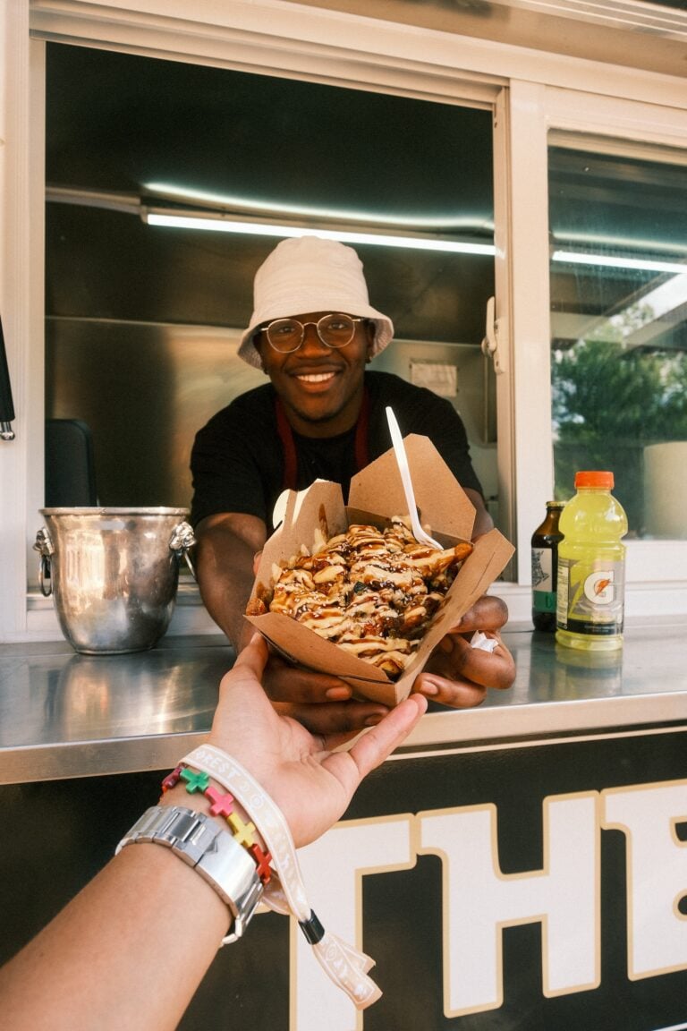 Personne souriante dans un camion-restaurant tendant une barquette de frites nappées de sauce à un festivalier
