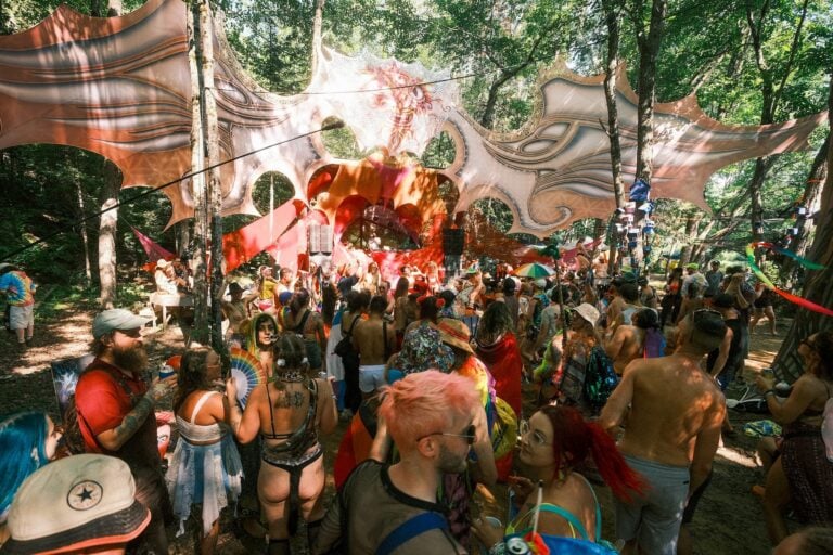 Foule qui danse en plein jour sous une structure de tissu orange et crème tendue entre les arbres, cabine de DJ au fond