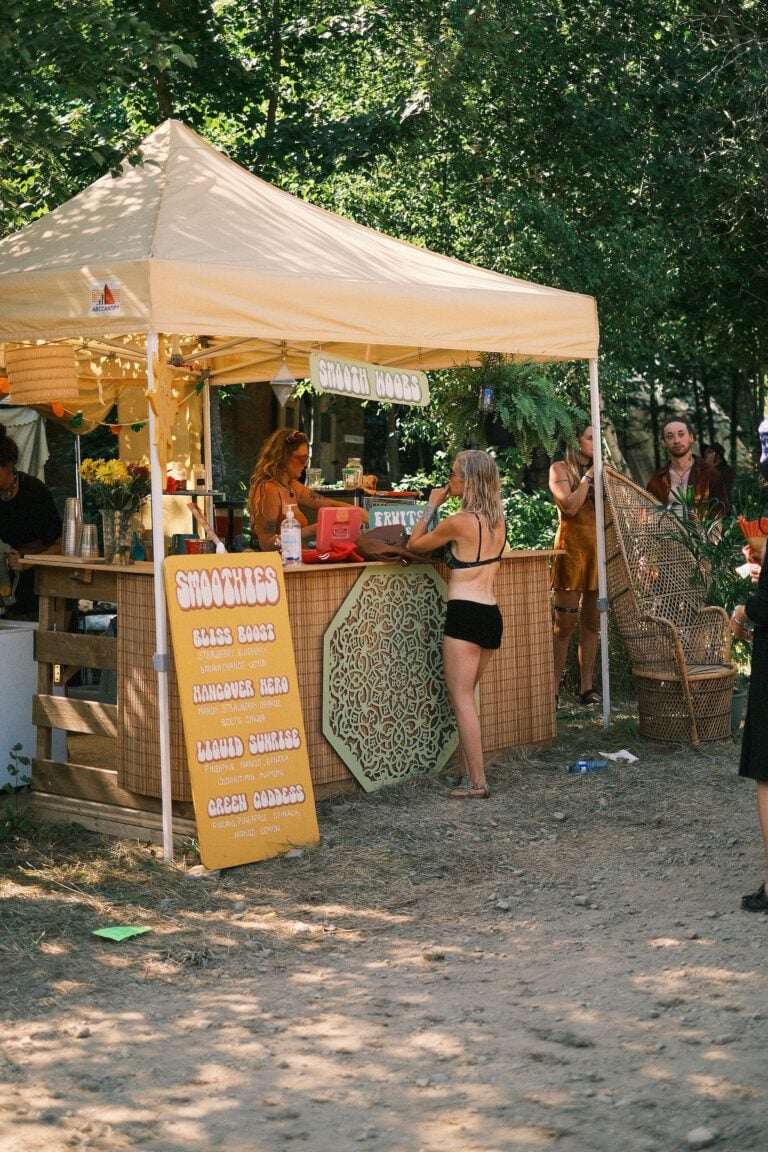Comptoir à smoothies sous un abri blanc en forêt, avec un menu jaune à l'avant et un fauteuil en rotin à côté
