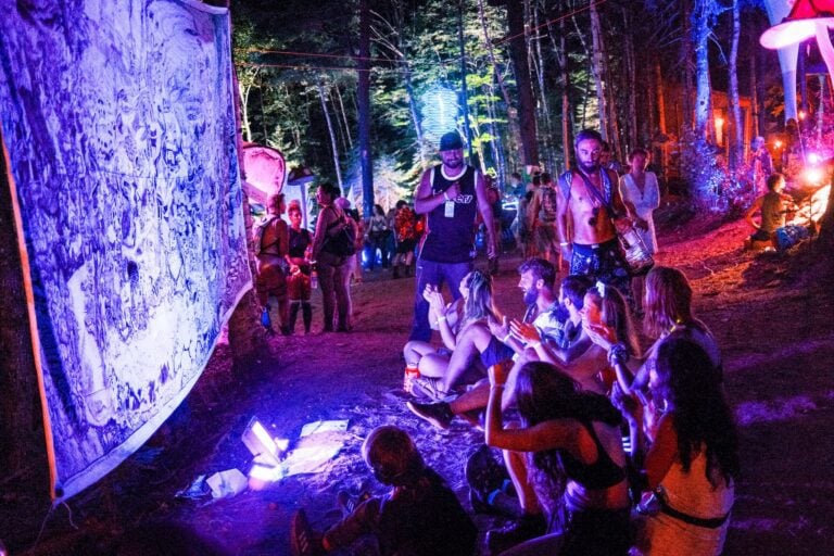 Des festivaliers assis la nuit sur un sentier forestier devant une grande murale dessinée à la main, éclairée en violet