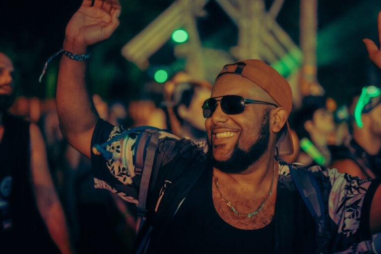 Festivalier souriant, lunettes de soleil et casquette à l'envers, lève le bras dans la foule de nuit sous des lumières vertes