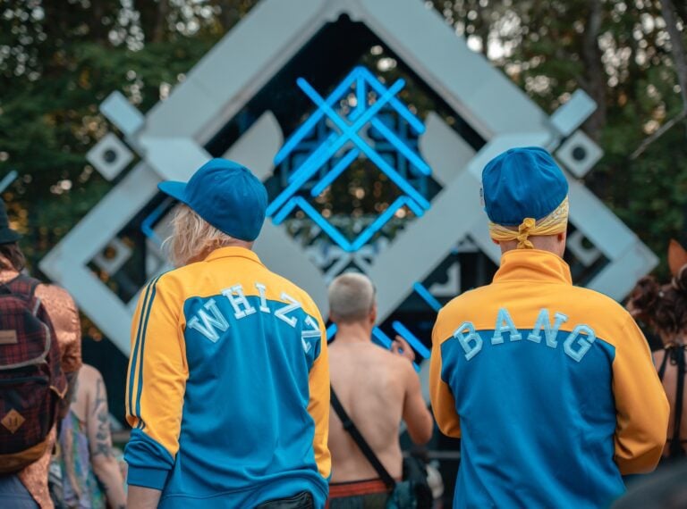Deux festivaliers en vestes bleu et jaune assorties, marquées WHIZZ et BANG, face à une scène blanche aux néons bleus