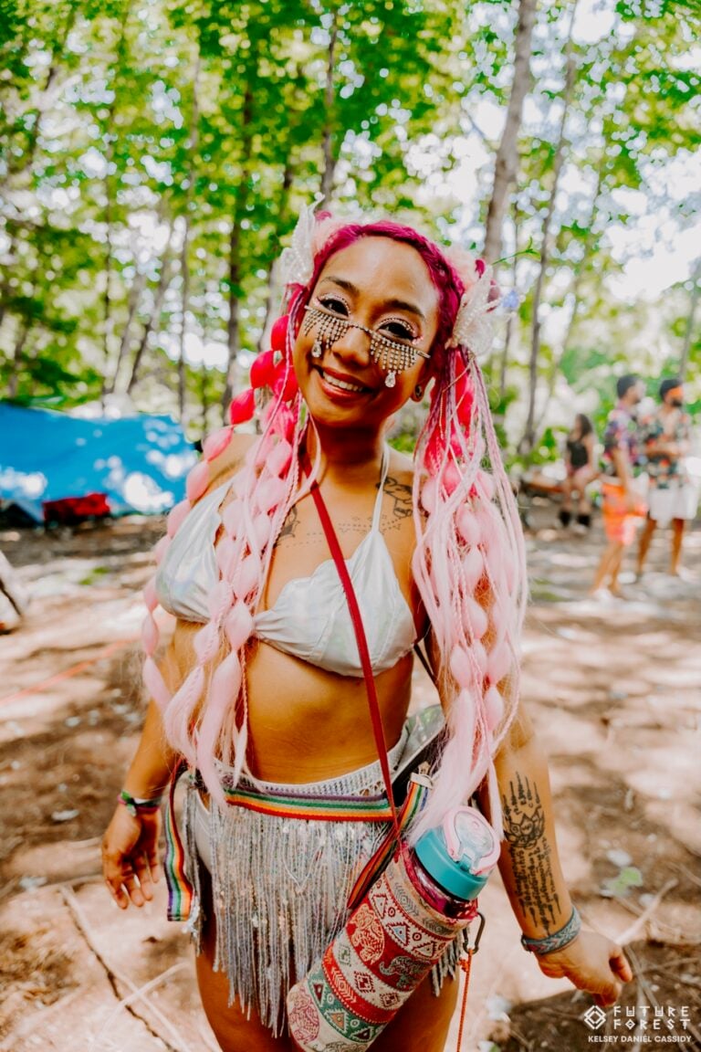 Une festivalière souriante aux longues tresses roses, bijoux de visage et jupe à franges argentée, dans un camping en forêt