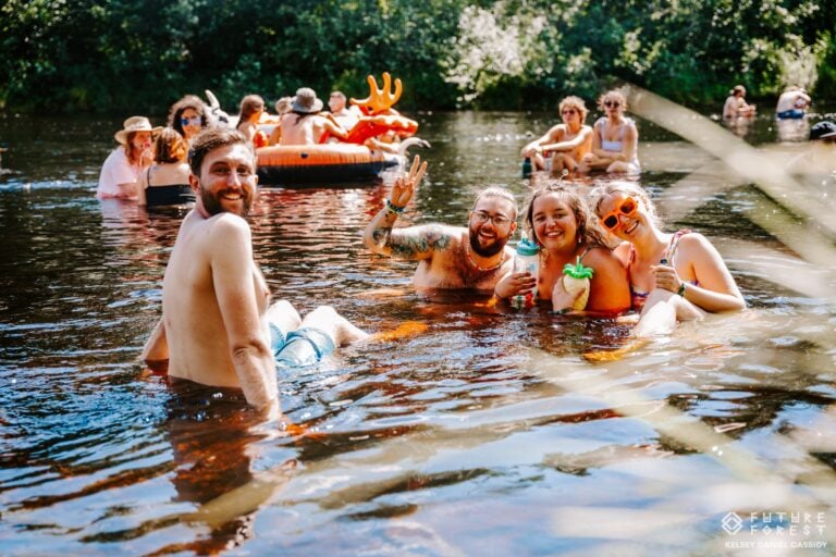 Des amis assis dans une rivière ensoleillée sourient à l'objectif, l'un fait le signe de paix, une bouée orange derrière eux