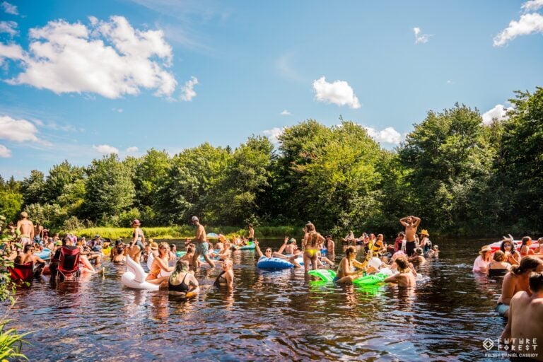Des dizaines de festivaliers dans une rivière par une journée ensoleillée, avec bouées et chaises pliantes, devant la forêt