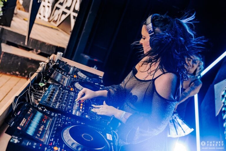 Une DJ aux longs cheveux foncés en mouvement mixe aux platines dans un éclat de lumière bleue, une autre personne derrière