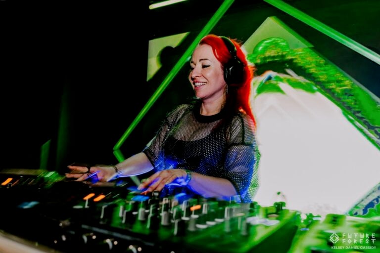 Une DJ aux cheveux roux, casque sur les oreilles, sourit en mixant sous un éclairage de scène vert, devant une projection