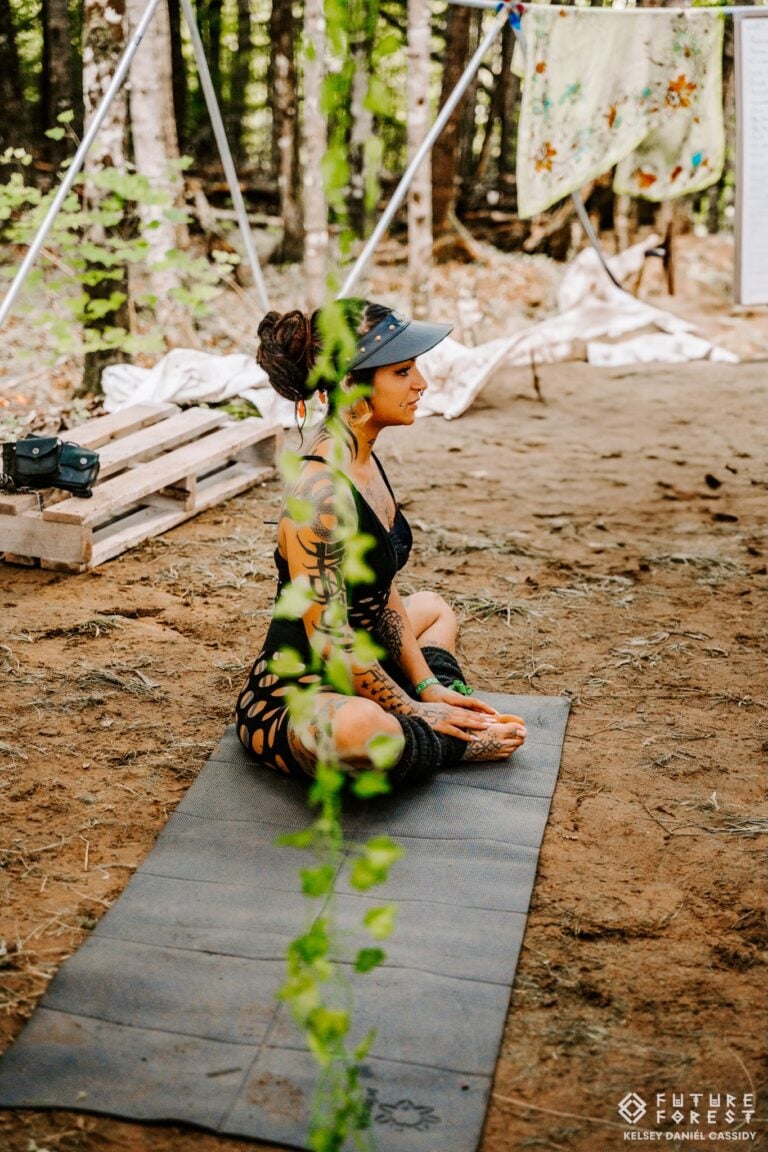 Une femme assise en tailleur sur un tapis de yoga, à même la terre d'une clairière en forêt, une liane verte au premier plan