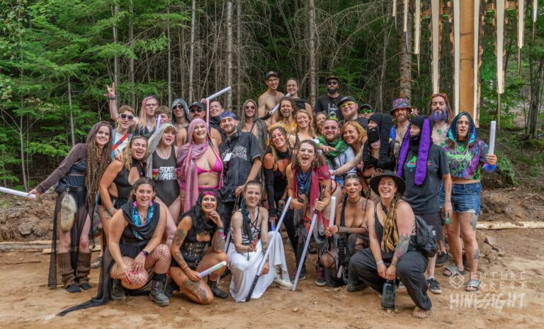 Grand groupe de festivaliers costumés posant ensemble dans une clairière de terre devant des conifères, certains tenant des bâtons lumineux