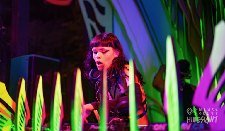 DJ à la frange rouge foncé et au casque autour du cou, penché sur les platines, entouré de feuilles décoratives vert néon et rose
