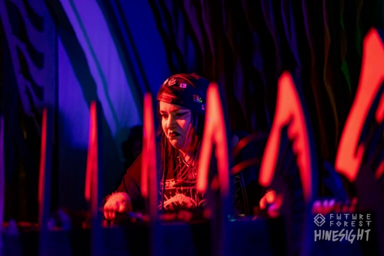 DJ au foulard rouge et aux vêtements sombres devant la table de mixage, éclairé en violet, derrière des formes pointues orange lumineuses