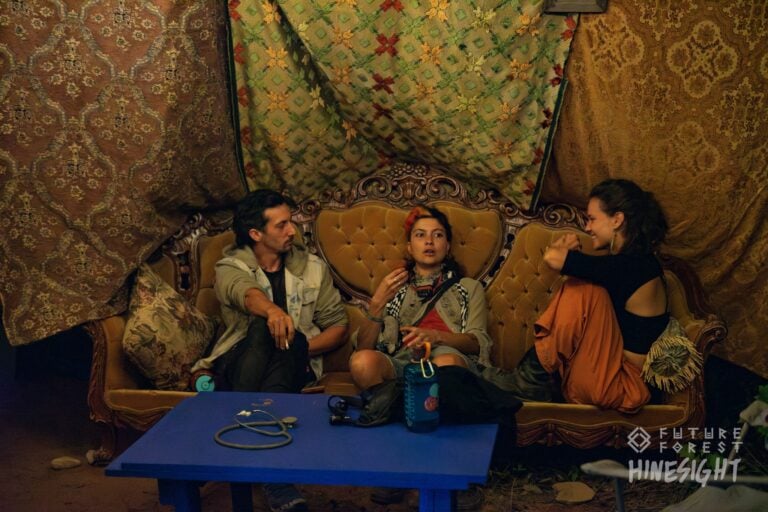 Trois festivaliers qui discutent sur un canapé ancien en velours doré sous des tissus à motifs suspendus, devant une table bleue
