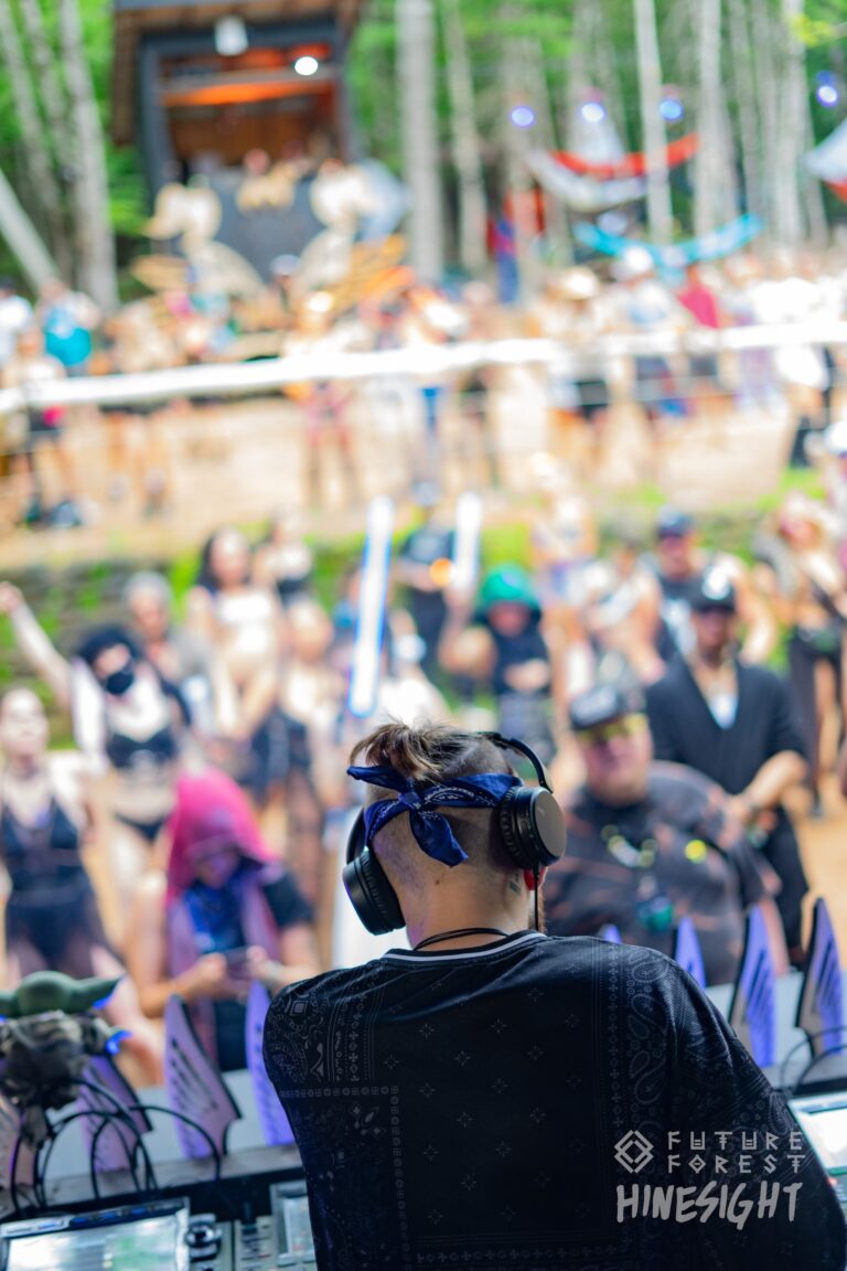DJ au casque et au bandana bleu vu de dos, face à une foule qui danse en plein jour les bras levés près d'une scène en forêt