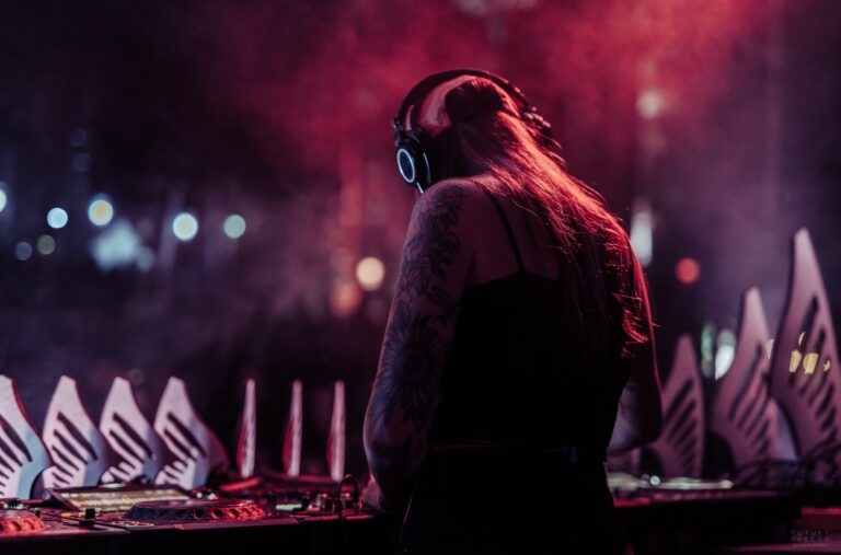 DJ de dos avec casque d'écoute et longs cheveux, bras tatoué, derrière les platines dans une brume rouge, avec des découpes de bois en feuilles