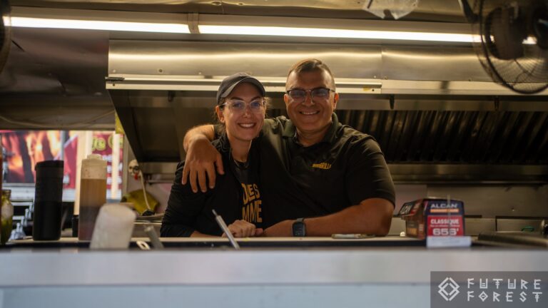 Deux personnes souriantes, bras autour des épaules, derrière le comptoir d'une roulotte de cuisine, avec une hotte en acier au-dessus