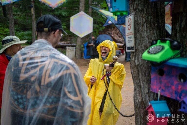 Personne qui rit en poncho de pluie jaune, tenant le combiné d'un téléphone vert à cadran près d'un arbre couvert de nichoirs peints