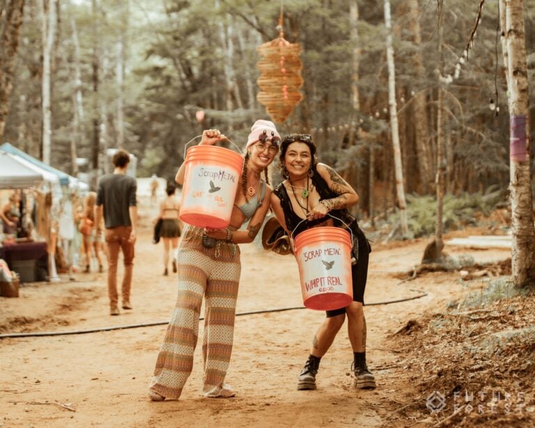 Deux festivaliers souriants tenant des seaux orange marqués Scrap Metal sur un chemin de terre bordé de tentes et d'arbres