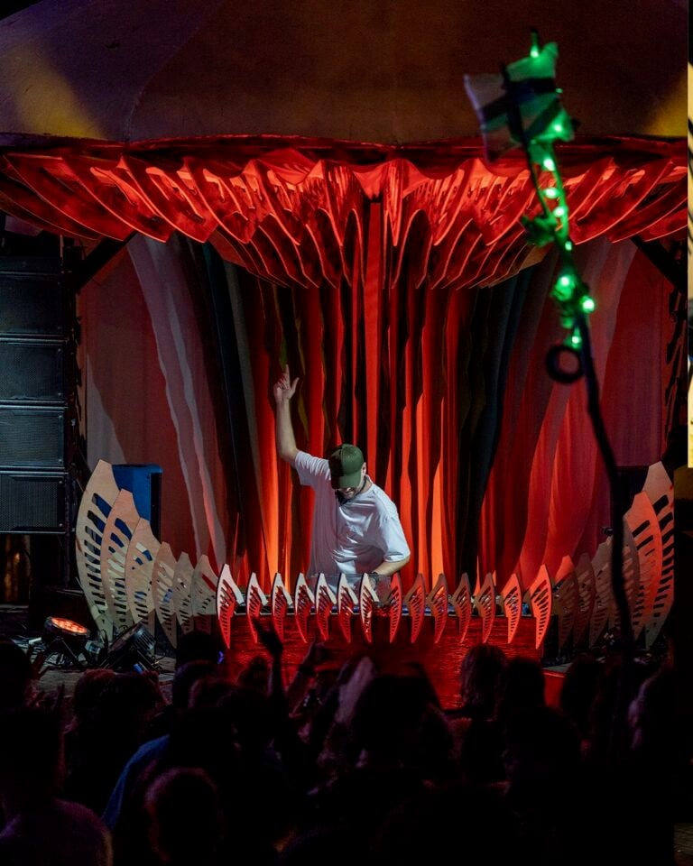 DJ en casquette verte et t-shirt blanc, une main levée, sur une scène aux rideaux plissés rouges et ailerons blancs, foule devant
