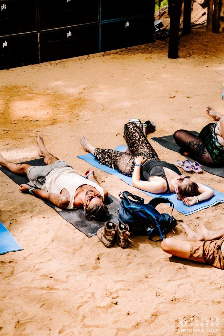 Festivaliers allongés sur des tapis de yoga dans le sable devant les enceintes d'une scène, avec bottes et sac à dos à côté