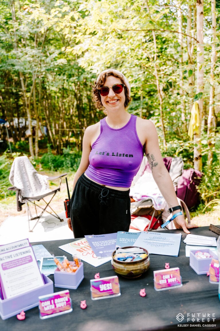 Personne souriante en camisole violette derrière une table de cartes sur le consentement et de dépliants en forêt