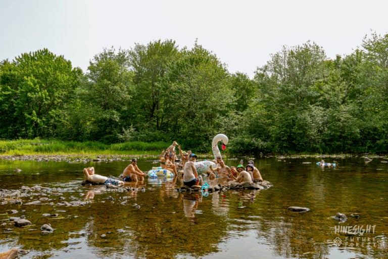 Groupe détendu dans une rivière peu profonde et rocailleuse, avec des bouées et un cygne gonflable, devant des arbres