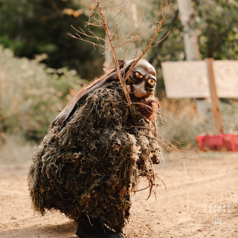 Artiste costumé en habit de mousse hirsute et masque sculpté aux yeux orange, tenant une branche nue sur un chemin de terre
