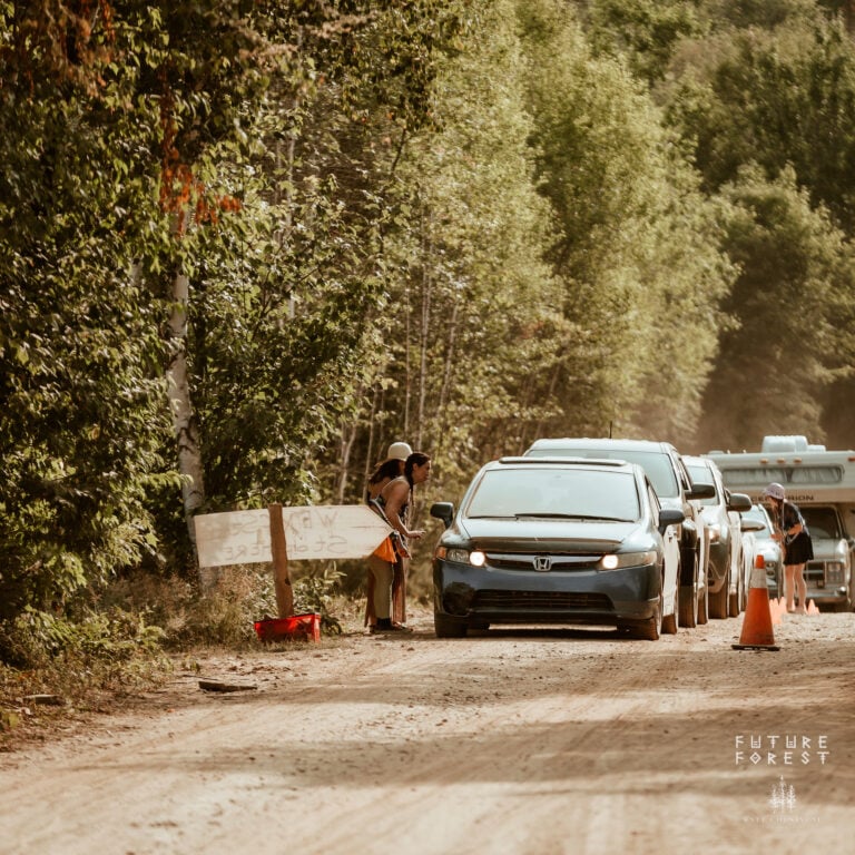 Voitures et autocaravane en file sur un chemin de terre, deux personnes près d'une pancarte peinte accueillent la première