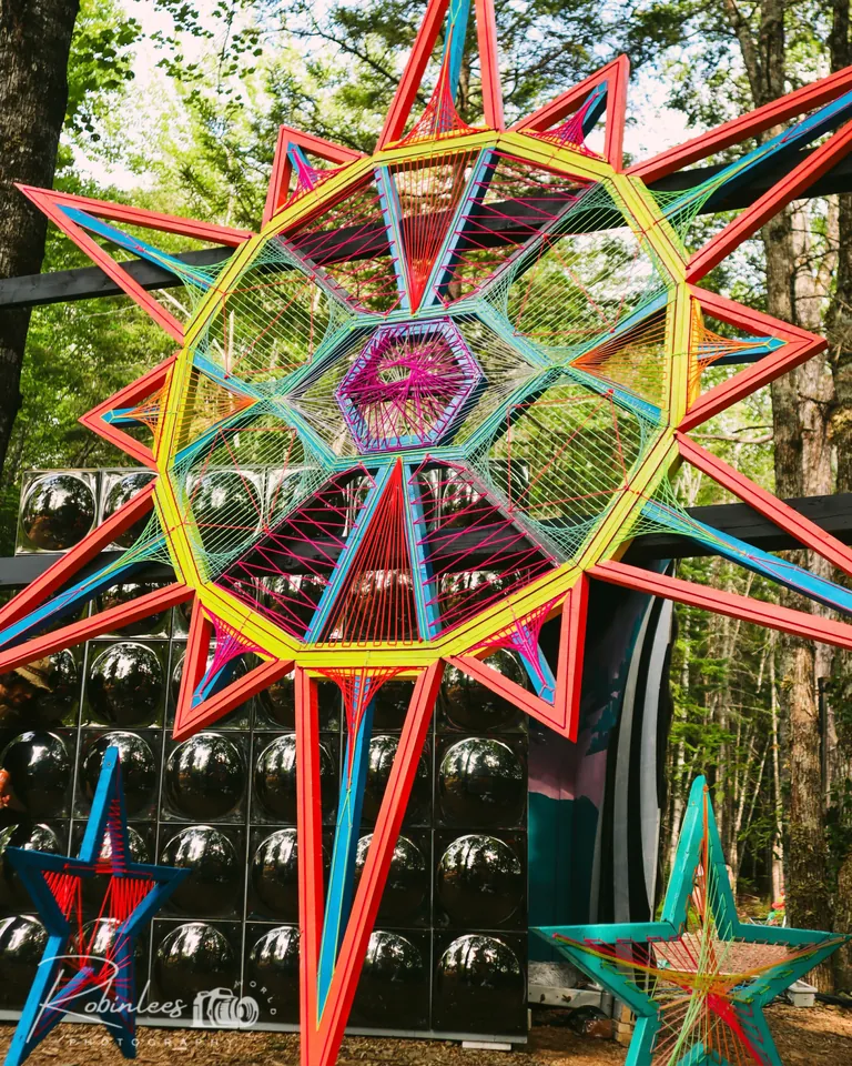 Étoile géante en art filaire rouge, jaune et turquoise en forêt, devant un mur de sphères miroirs et de petites étoiles